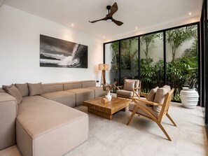 A modern living room with a beige sofa, a wooden coffee table, and two chairs. There's a large window with plants visible outside. - Elegant 5BR Villa Retreat Steps From Canggu Beach (Munggu)