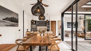 A modern dining area with a wooden table and chairs, a ceiling fan, and a view through a glass door to an outdoor space. - Stylish 5BR Beach Villa, 200m in Boho Canggu (Munggu)