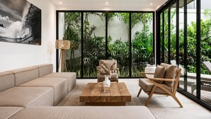 A modern living room with a sofa, coffee table, and chairs, large windows with a view of greenery, and a ceiling fan. - Stylish 5BR Beach Villa, 200m in Boho Canggu (Munggu)