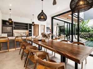 A modern dining area with a long wooden table and chairs, a kitchen with a built-in oven, and a large glass door leading to a garden. - Elegant 5BR Villa Retreat Steps From Canggu Beach (Munggu)
