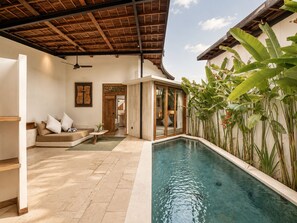 Un área junto a la piscina con techo de madera, un sofá con cojines, una mesita con un florero y una puerta de vidrio que da a una habitación.