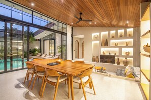 A modern dining area with a wooden table and chairs, a ceiling fan, and a pool view. - Luxurious 5BR Boho Sanctuary, Near Bingin Beach (Pecatu)
