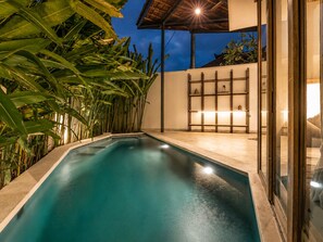 A swimming pool with a tiled deck, surrounded by lush greenery and a modern outdoor seating area with a glass partition. - Lush 1-br Villa With Pool, 4 Mins to Echo Beach, (Canggu)