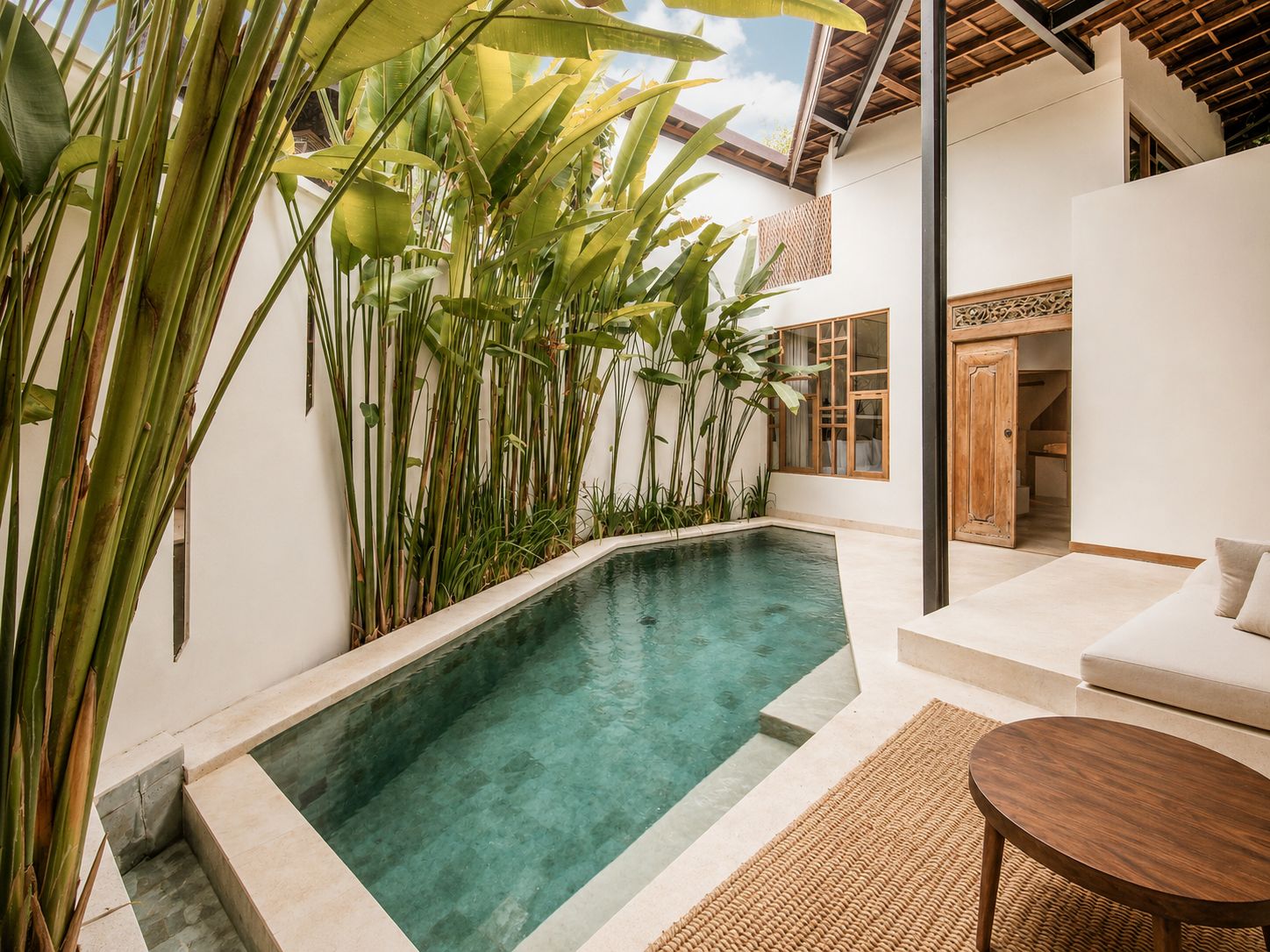 A pool with clear water, surrounded by tall green plants, a white wall, and a wooden door.