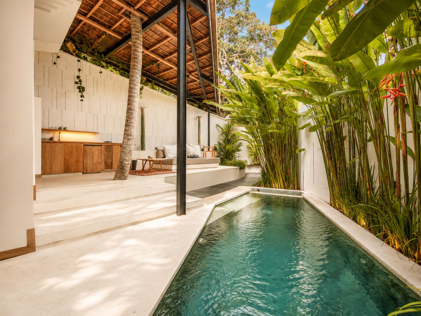 A modern outdoor pool area with a stone deck, a wooden bench, and lush tropical plants.