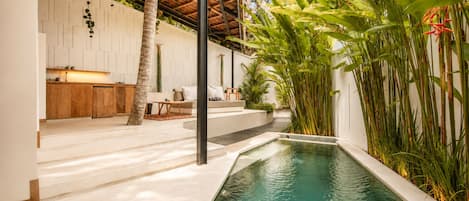 A modern outdoor pool area with a stone deck, a wooden bench, and lush tropical plants.