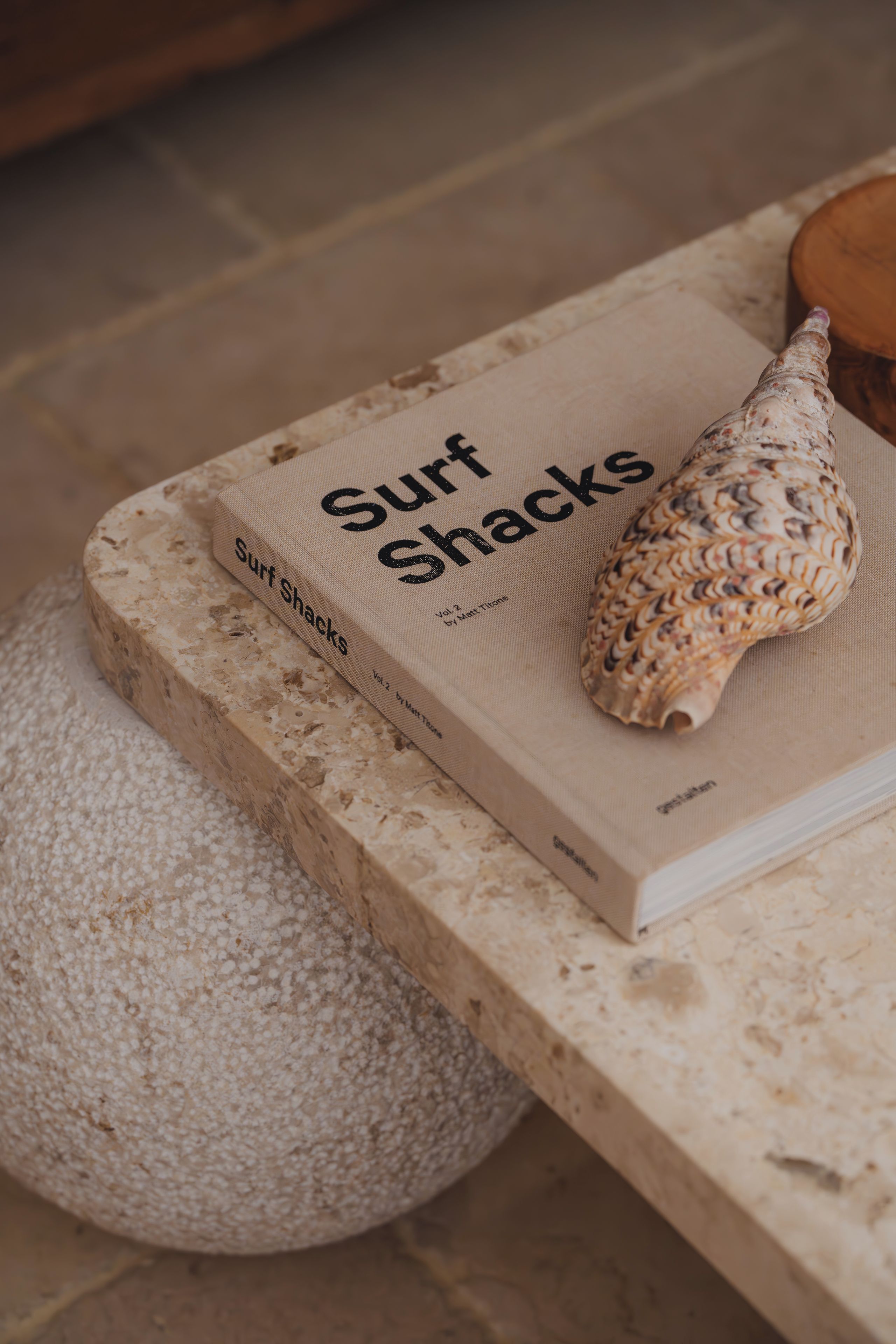 A book titled 'Surf Shacks' on a textured surface with a seashell beside it.