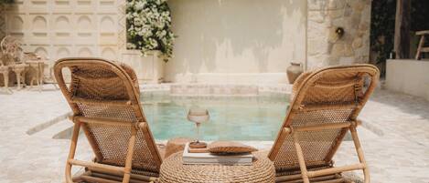 Two wicker lounge chairs with a book and a glass on a woven mat, overlooking a pool with a stone wall and floral decorations in the background.