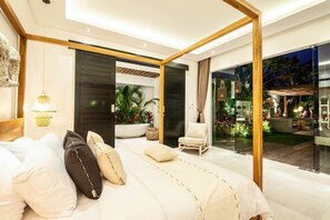 Une chambre moderne avec un grand lit, un fauteuil et une vue sur un jardin luxuriant, visible à travers des portes vitrées.