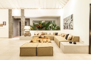 A spacious living area with a large L-shaped sectional sofa, a coffee table with a natural wood top, and a side table. There is a lamp with a white shade and two wicker baskets on a console table. The room features a stone accent wall, a large window with a view of greenery, and a framed black and white photograph on the wall. - Modern 4 BR Villa 400m to the Beach in Canggu (Canggu)