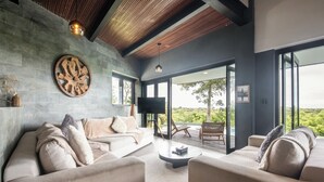 A modern living room with a large sofa, a coffee table, and a television. There is a large window with a view of greenery outside. - Elegant 6BR Clifftop Villa Near Bingin (Pecatu)