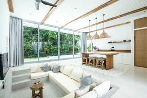 Una sala moderna con un amplio sofá modular, una mesa de centro de madera y una zona de cocina con encimera de madera y lámparas colgantes.