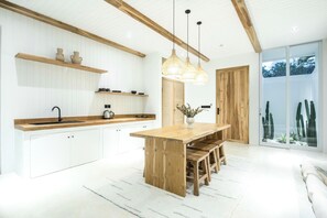 Una cocina moderna con armarios blancos, vigas de madera, una isla central con banquetas, iluminación colgante y vistas a cactus a través de puertas de vidrio.