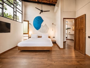 Quarto com cama, ventilador de teto, televisão e uma grande janela com plantas.