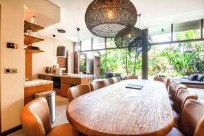 A modern kitchen with a large island, a dining area with a round table and chairs, and large windows offering a view of greenery. - Luxury 6BR Canggu 400m to Finns Beach (Canggu)