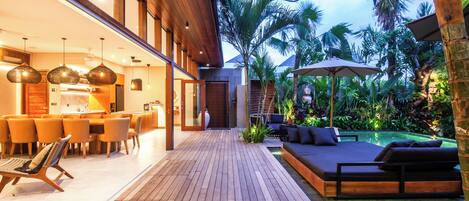 A wooden deck with a dining area, a pool, and a lounge with a view of the surrounding greenery.