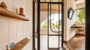 A modern interior with a wooden bench, a shelf with decorative items, a television, and a view of greenery outside. - Modern 1-br Loft With Art Deco Vibe Shared Pool (Canggu)