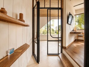 A modern interior with a wooden bench, a shelf with decorative items, a television, and a view of greenery outside. - Modern 1-br Loft With Art Deco Style Shared Pool (Canggu)