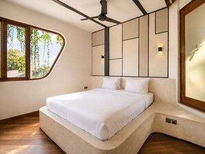 A modern bedroom with a curved bed, a ceiling fan, a large window with a view of greenery, and a wall-mounted light fixture. - Modern 1-br Loft With Art Deco Vibe Shared Pool (Canggu)