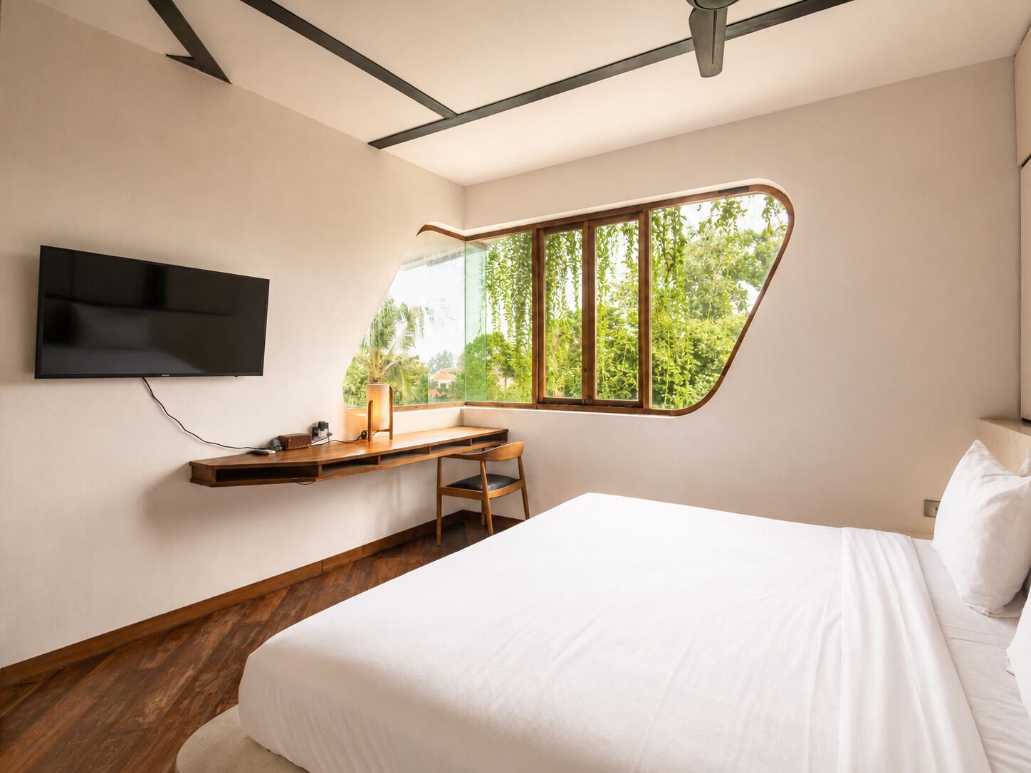 A bedroom with a bed, a desk, a chair, a TV mounted on the wall, and a window with a view of greenery.