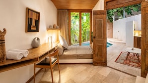 A room with a wooden desk, a chair, a sofa, a painting, and a view of a pool and greenery outside. - Chic 1-br Villa With Pool, 4 Mins to Echo Beach (Canggu)