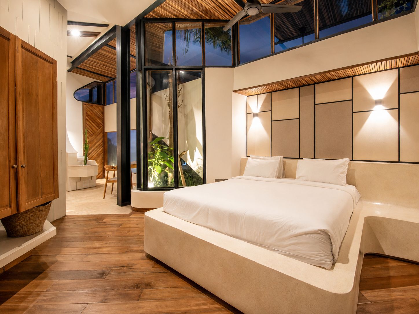 A modern bedroom with a large bed, wooden flooring, and a view of the outdoors.