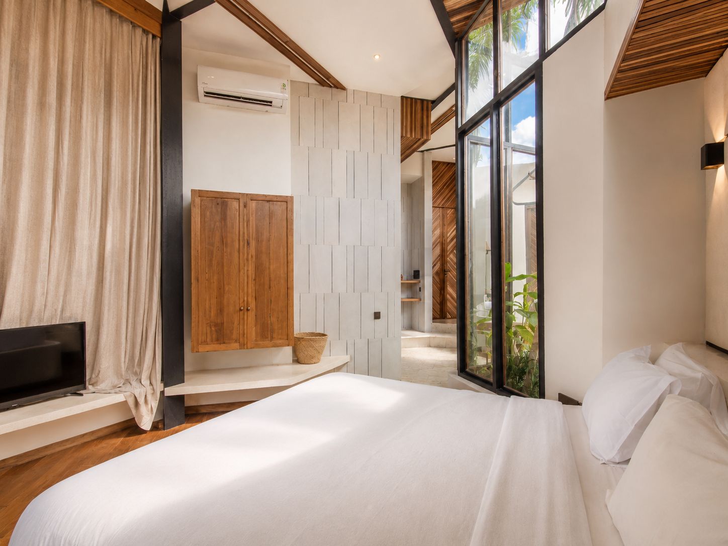 A modern bedroom with a bed, a TV, a wardrobe, and a window with a view of greenery.