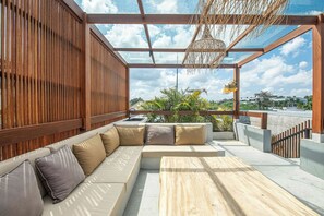 Ein moderner Außenbereich mit einer Holzbank und einer gepolsterten Couch, einem Holztisch sowie Blick auf Himmel und Grünpflanzen.