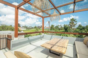 A spacious balcony with a wooden table and seating, large glass windows, and a view of greenery and buildings.