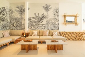 Una sala de estar moderna con un sofá color beige, dos mesas de centro de madera y un gran mural botánico en la pared.