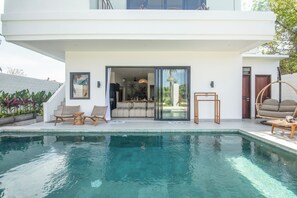Een modern huis met een zwembad, buitenmeubilair en een glazen deur die leidt naar een woonruimte.