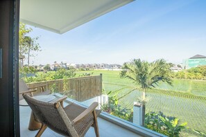 Een balkon met rieten stoelen en een tafel, met uitzicht op een rijstveld en woongebouwen.