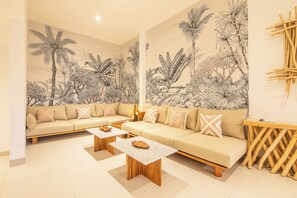 Een moderne woonkamer met een beige bank, twee houten salontafels en een groot muurdecor van tropische planten.