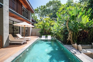 Une piscine entourée de chaises longues et d’un parasol, le tout baigné d’une végétation luxuriante et encadré par une architecture moderne.