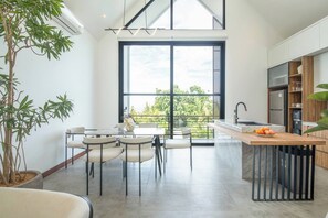 A modern kitchen with a large window, a dining area with chairs, and a potted plant. - Elegant Villa Sinara 2BR With Sea View (Kutuh)