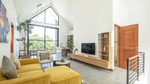Een ruime woonkamer met een hoge plafond, een gele bank, een televisie en een houten boekenkast.