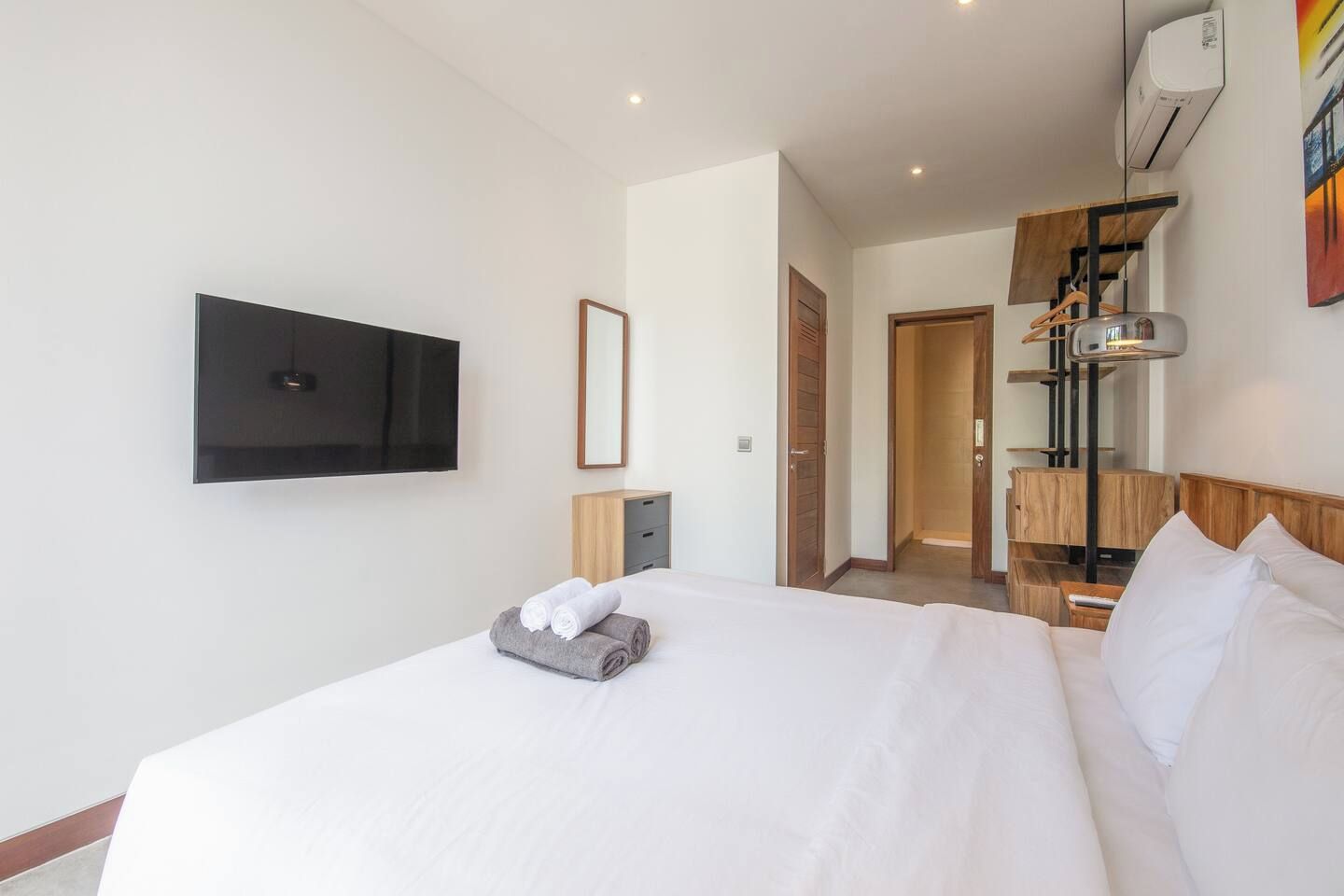 A hotel room with a large bed, a flat-screen TV mounted on the wall, a bedside table with a lamp, and a wooden headboard.