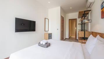 A hotel room with a large bed, a flat-screen TV mounted on the wall, a bedside table with a lamp, and a wooden headboard.