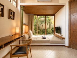 A room with a wooden desk, a chair, a sofa, a TV, and a window with a view of greenery.