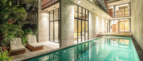 A modern indoor swimming pool with lounge chairs, concrete walls, and a wooden deck.