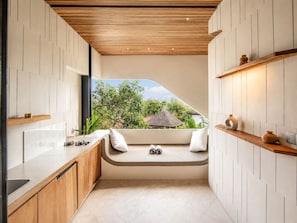 Une cuisine moderne dotée d’un banc intégré, d’étagères en bois et offrant une vue sur un paysage tropical.