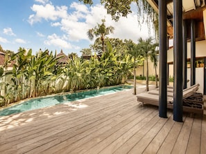 Une terrasse en bois avec une piscine, entourée de plantes tropicales et d’un bâtiment orné de colonnes.