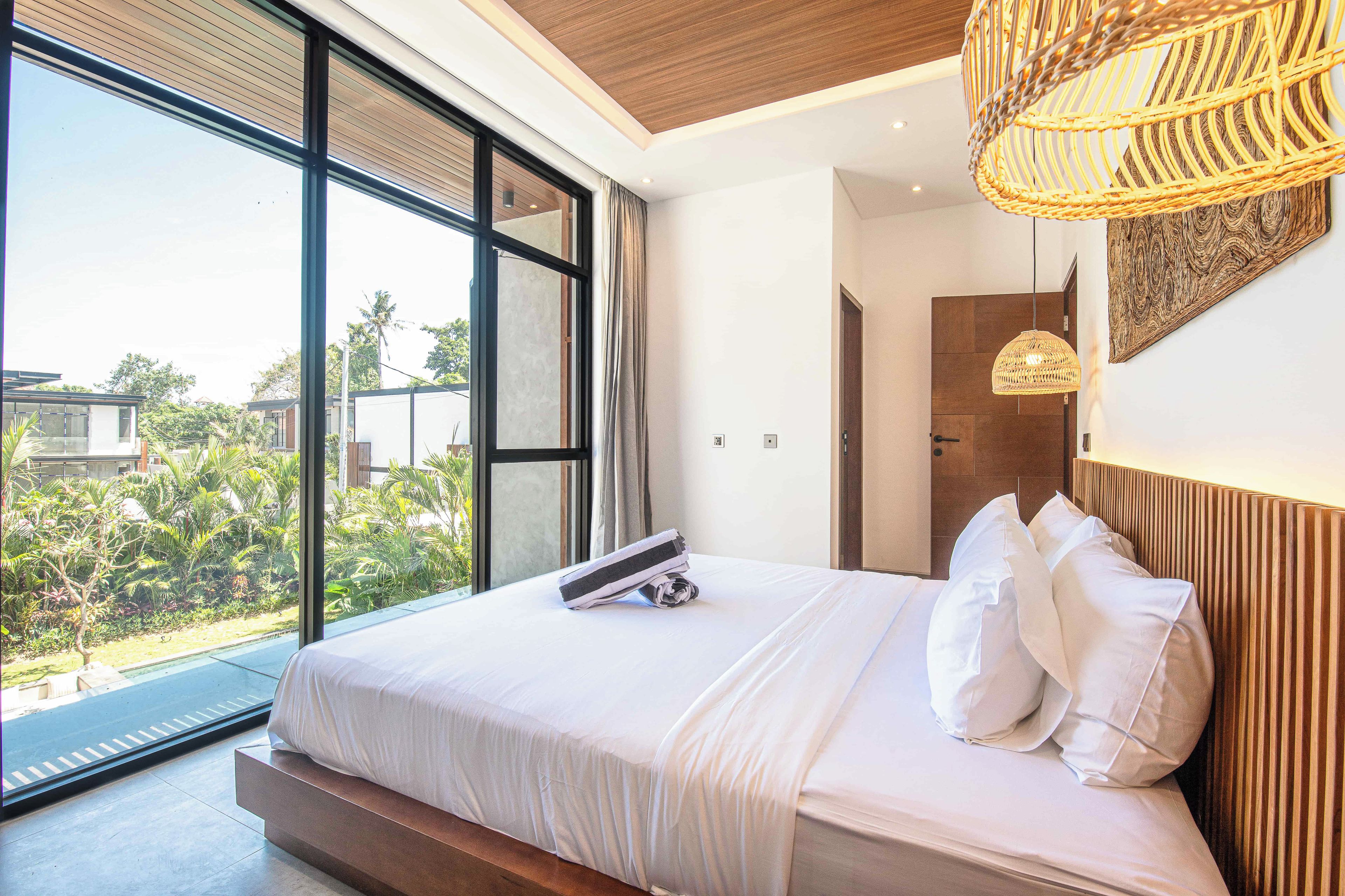 A modern bedroom with a large bed, a view of greenery outside, and a woven light fixture.