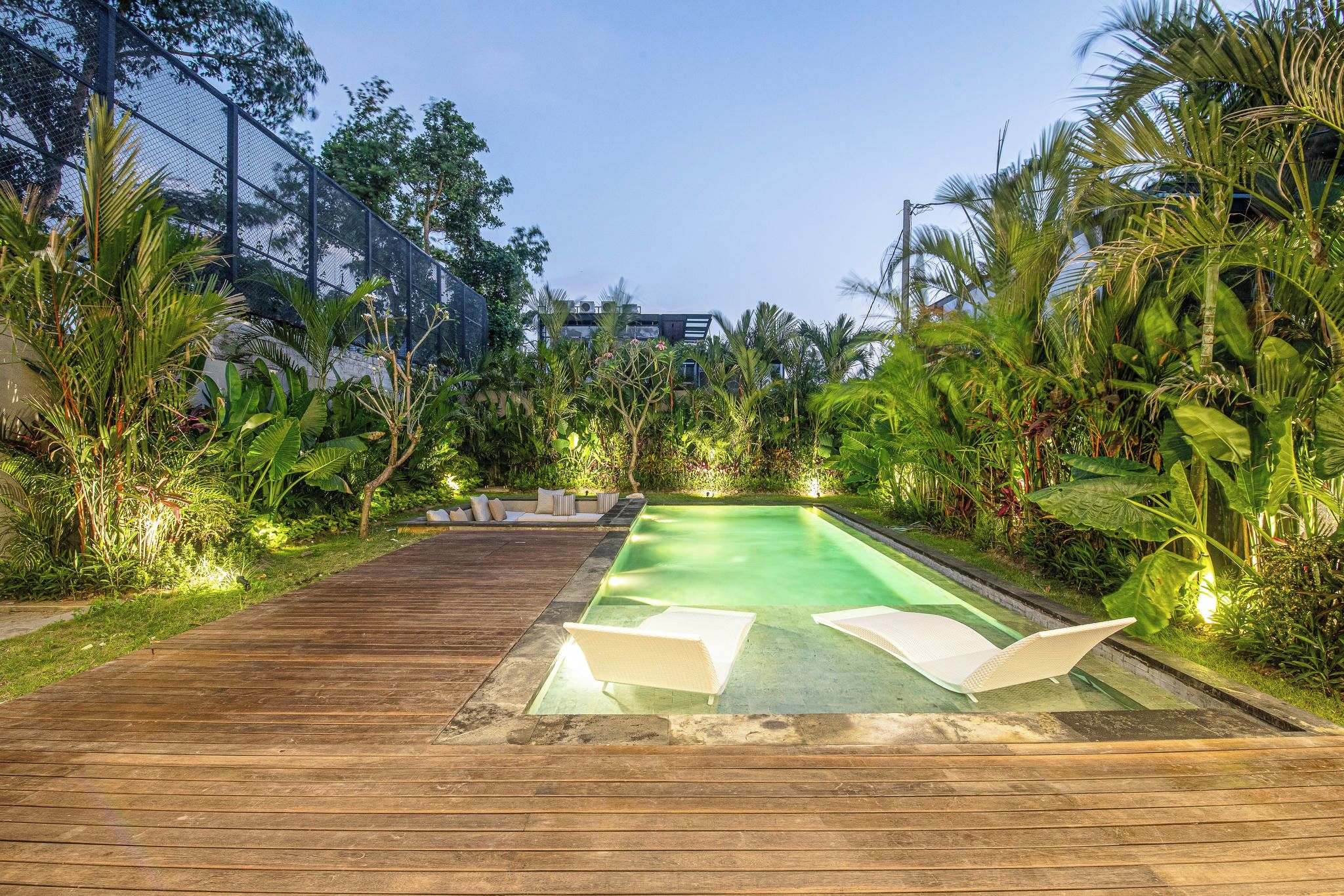 A rectangular swimming pool with two white lounge chairs, surrounded by lush greenery and a wooden deck.