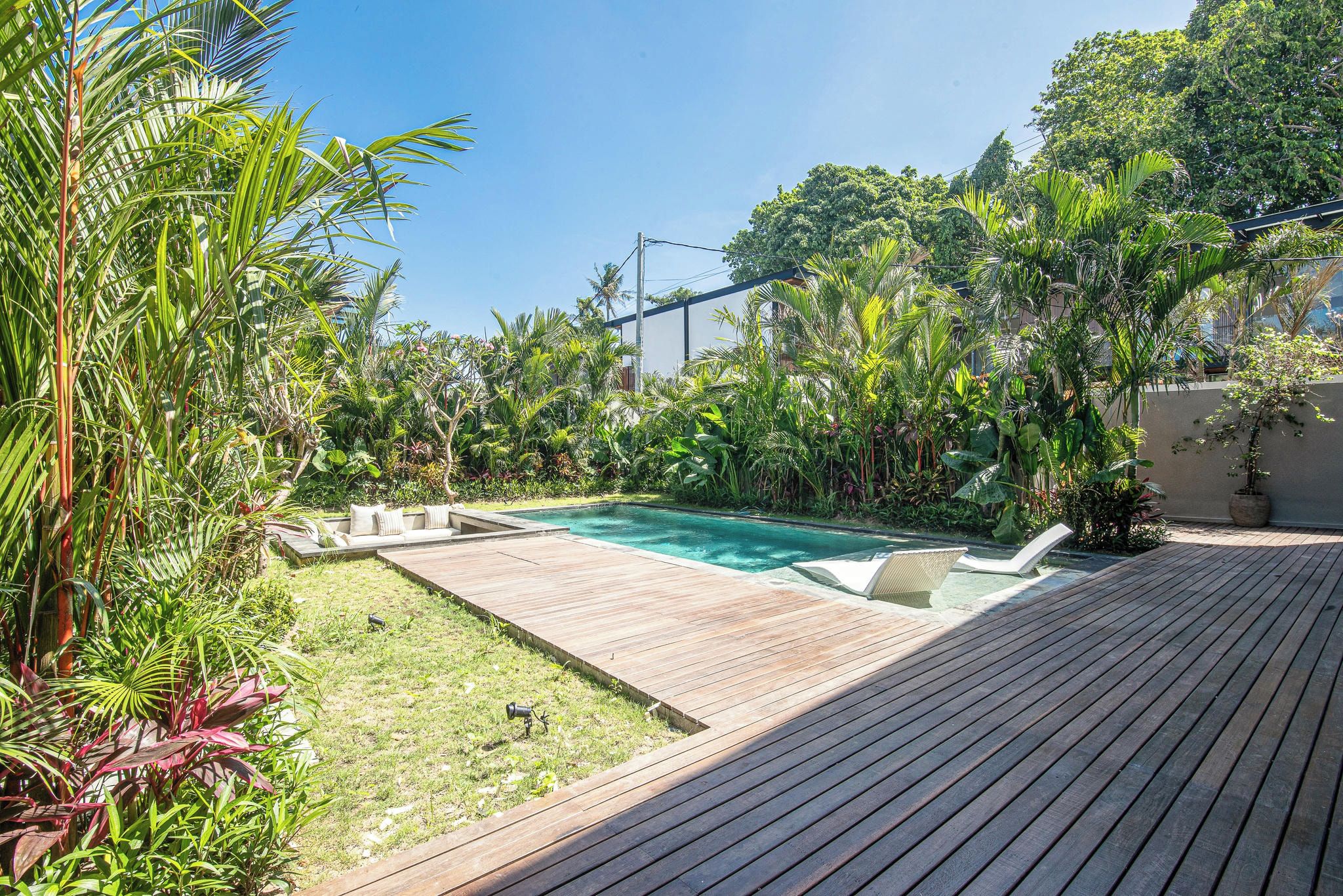 A wooden deck with a pool, surrounded by lush tropical plants and a clear blue sky.