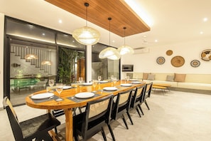 A modern dining area with a long wooden table set for six, surrounded by black chairs with woven backrests.