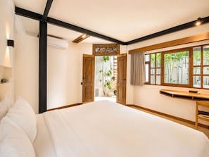 A bedroom with a large bed, a desk, a window with a view of greenery, and a door leading to another room.