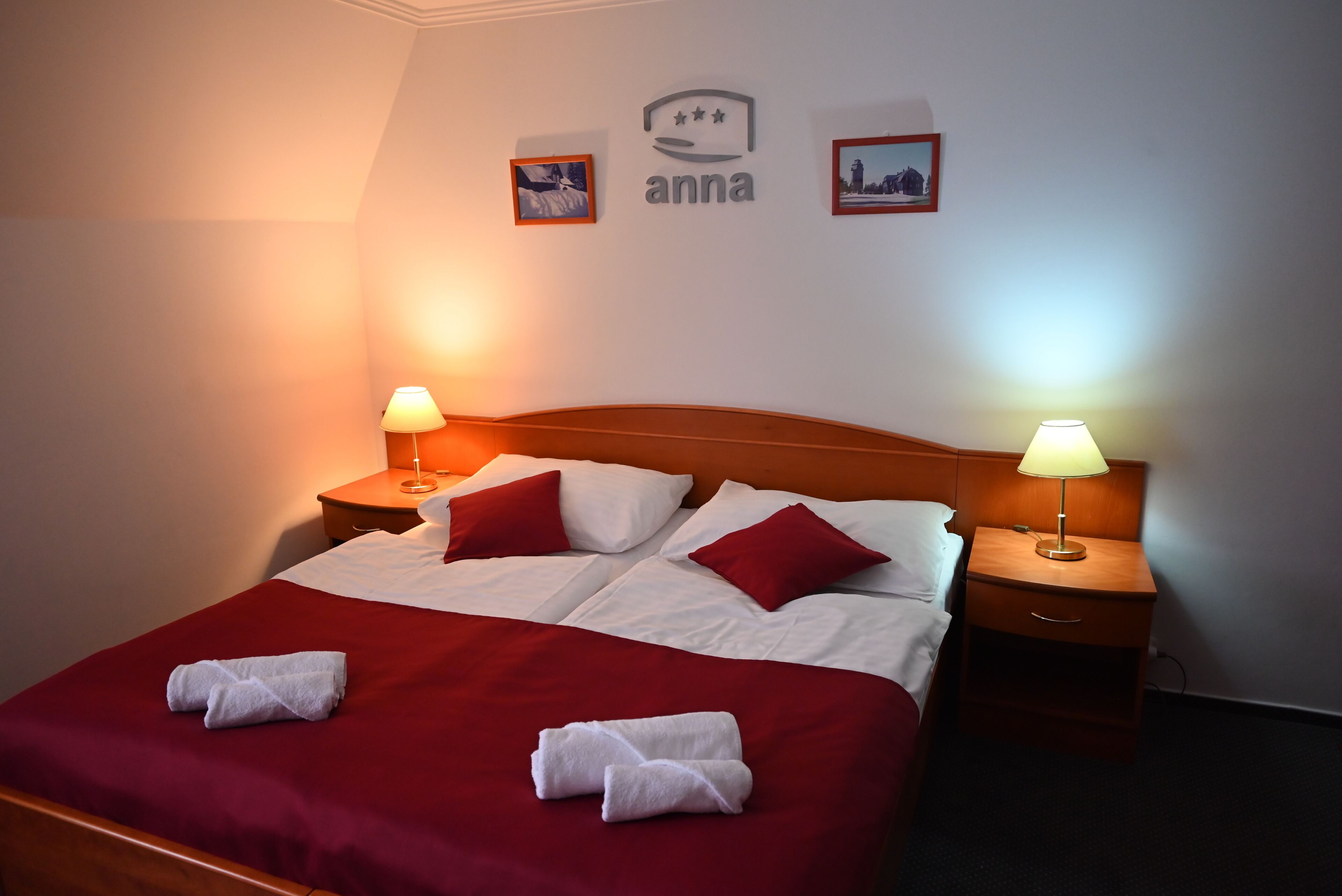 Una cama bien hecha con ropa de cama roja y blanca, dos lámparas de noche y una pared con cuadros enmarcados y un logotipo.