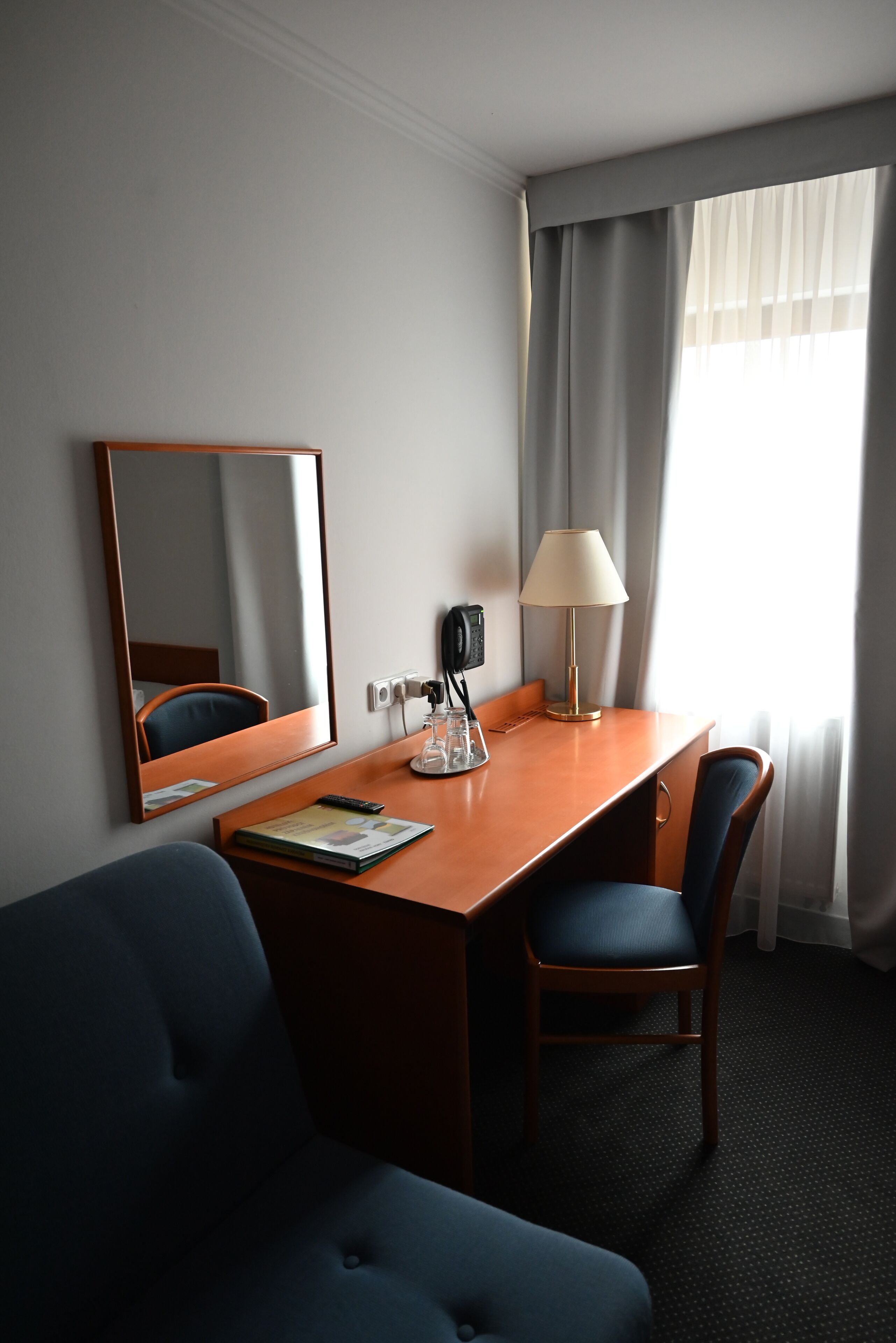A hotel room with a wooden desk, a chair, a lamp, a mirror, and a window with curtains.