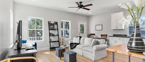 A spacious living room with a white sofa, a coffee table, and a TV. There is a bookshelf, a dining area, and a kitchen visible.